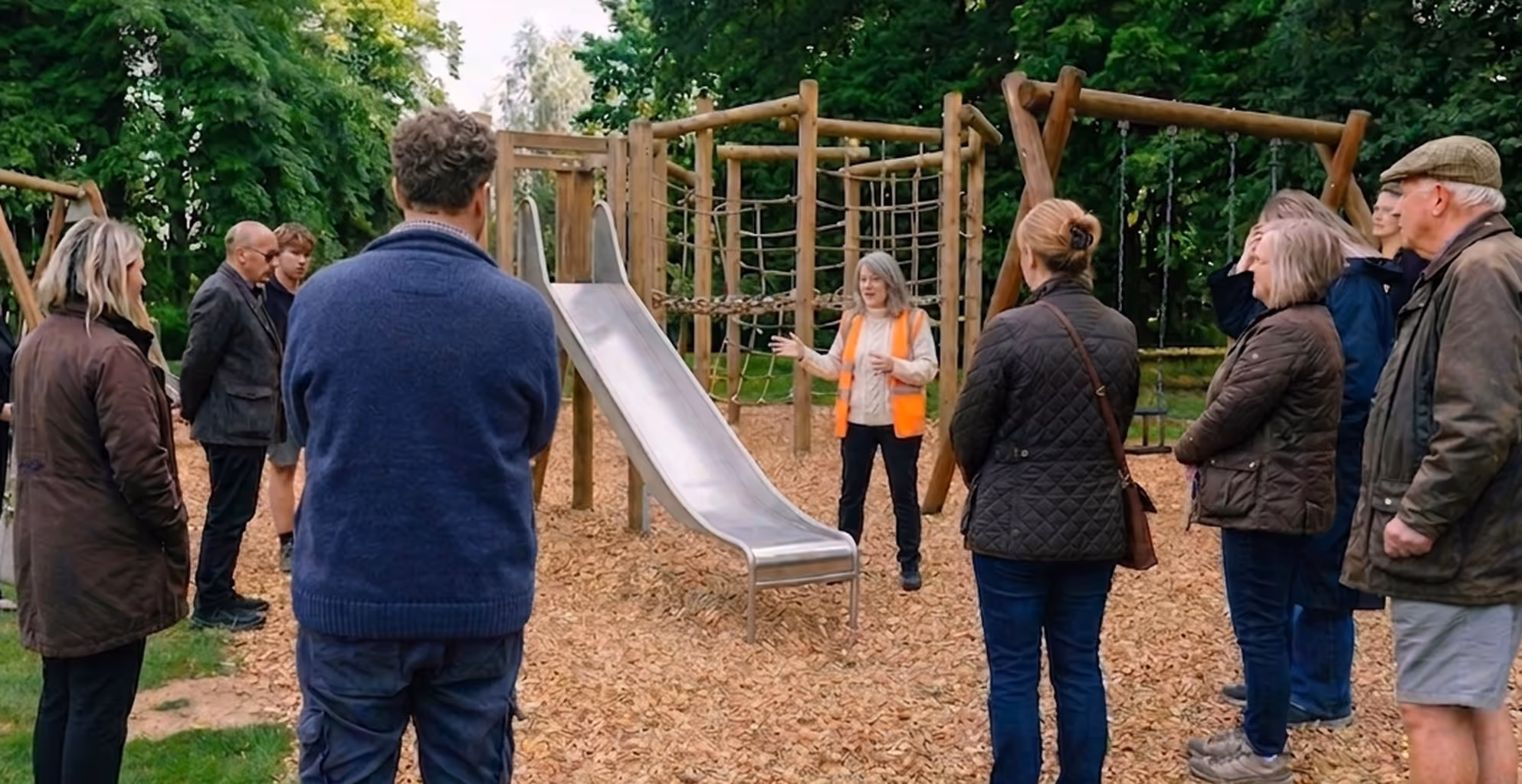 Group of adults listening to a woman in an orange vest explaining playground equipment with a slide and swings.