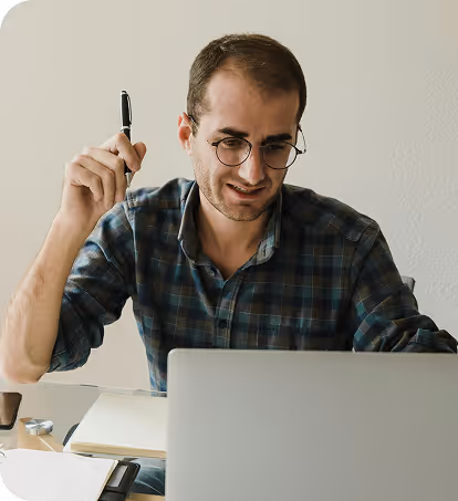 Man wearing glasses and a checkered shirt holding a pen, looking at a laptop screen while working.