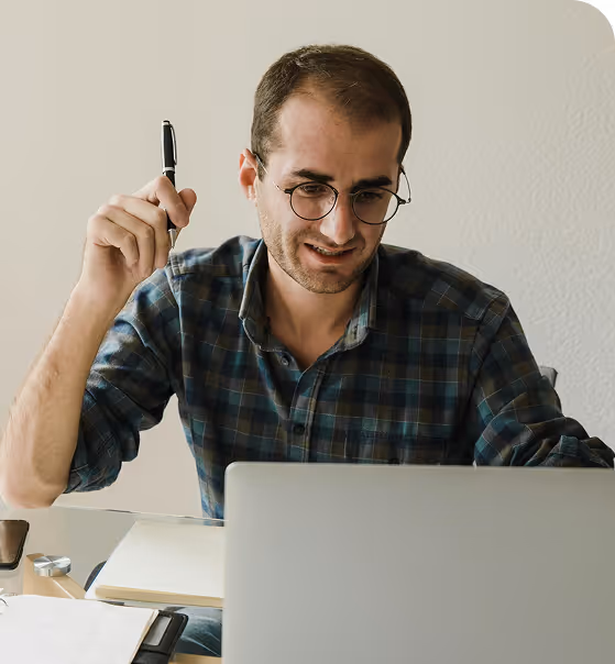 Woman in glasses sitting at desk writing notes while looking at a laptop screen with spreadsheet data.