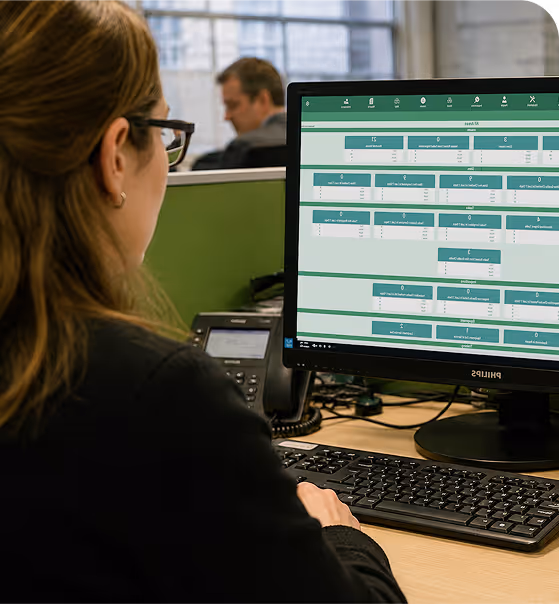 Woman in glasses sitting at desk writing notes while looking at a laptop screen with spreadsheet data.