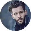 Man with dark hair and beard wearing a dark jacket against a blurred background.