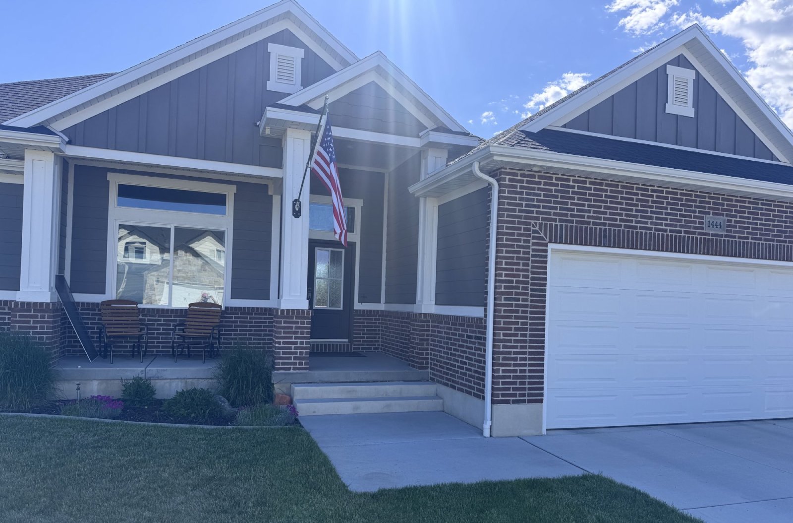 Front of a gray house with white trim, brick accents, American flag, and two chairs on the porch.