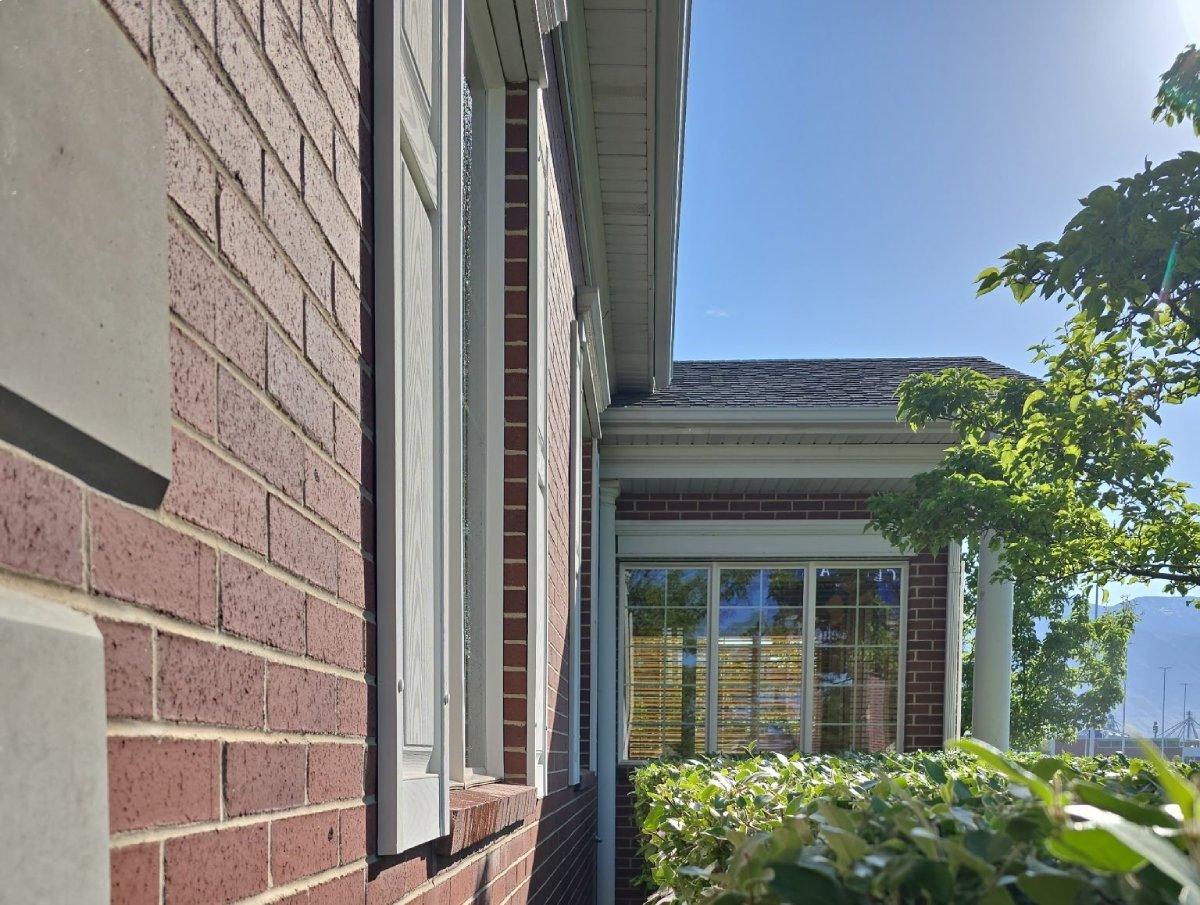 Side view of a brick house exterior with windows, green bushes, and a tree under a clear blue sky.