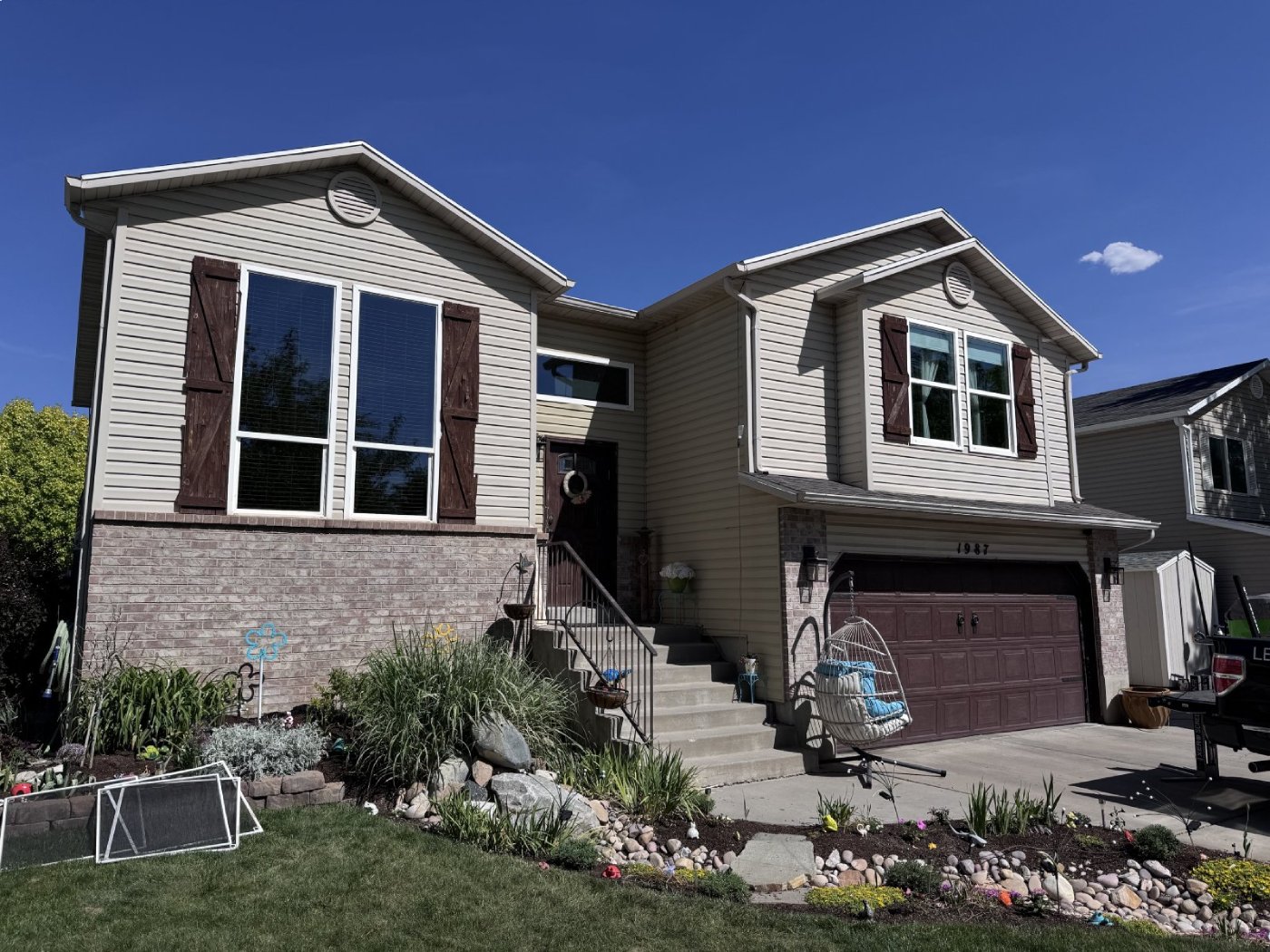 Two-story house with beige siding, brown shutters and garage door, a front porch, and a lawn garden.