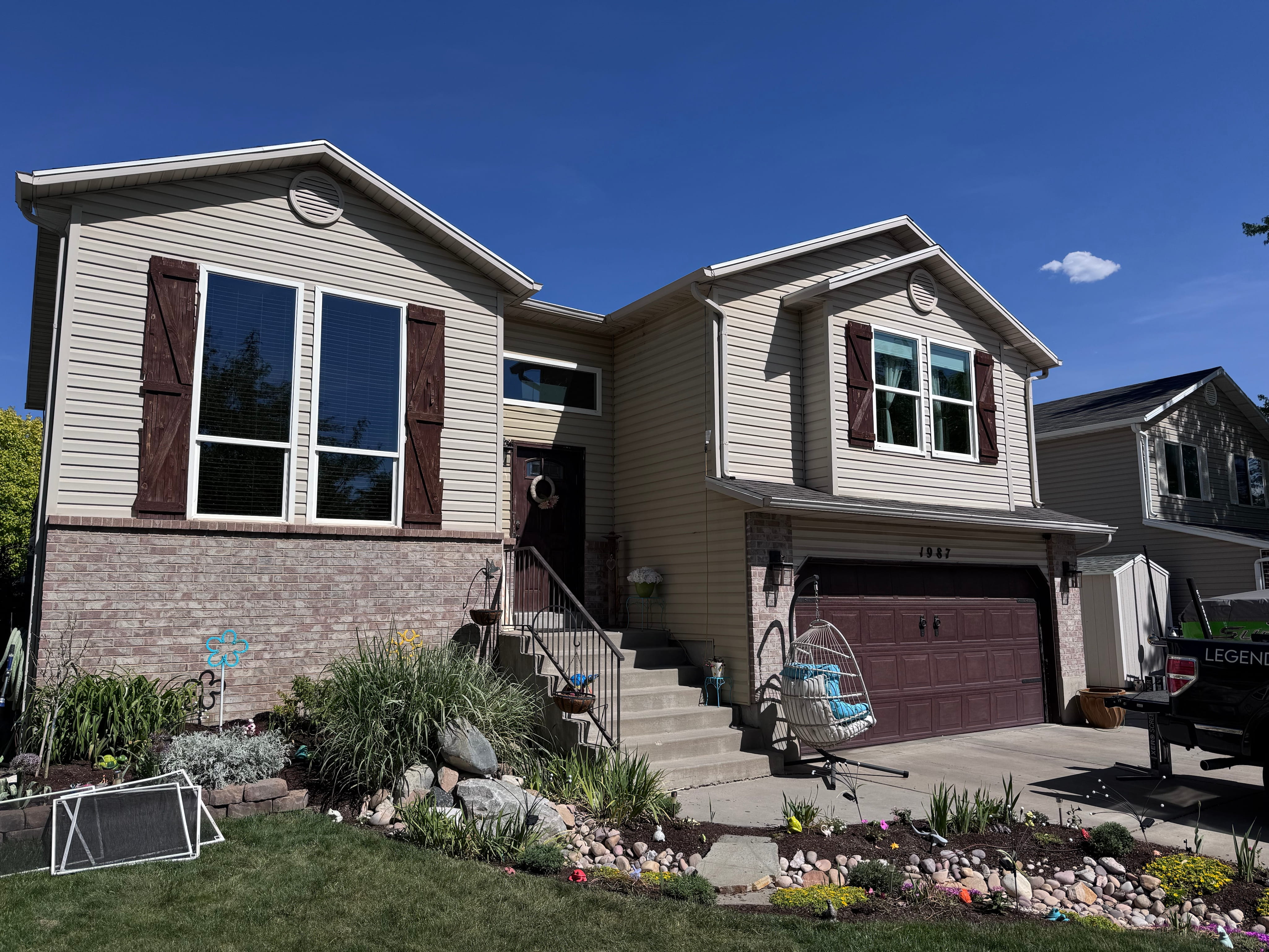 Two-story beige house with brick lower level, brown shutters, front steps, hanging chair, and garden.