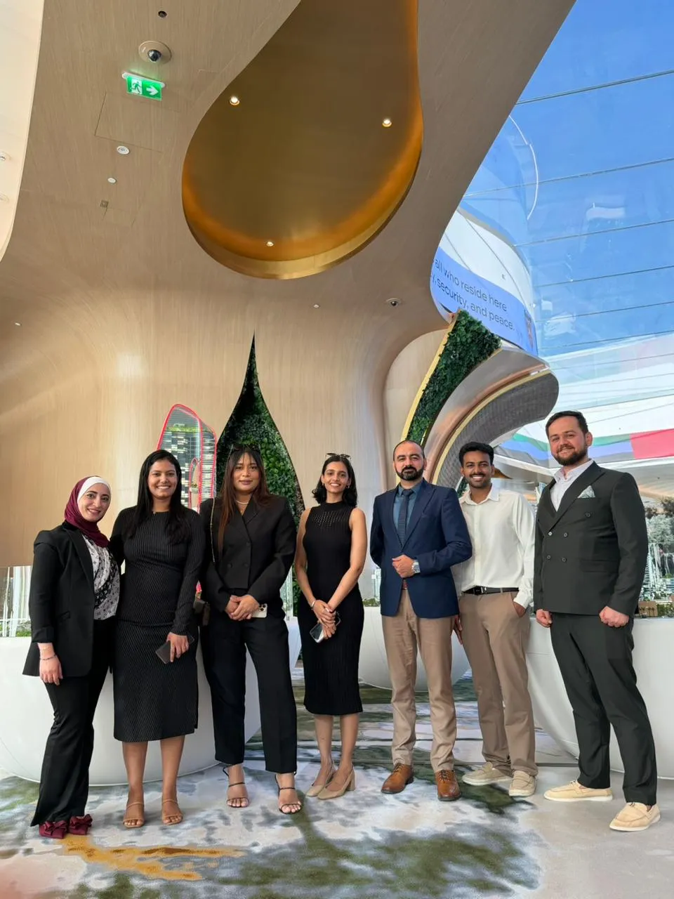 Group of seven professionally dressed people posing indoors under modern ceiling with gold and greenery accents.