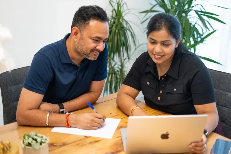 A man and woman work together at a wooden table, smiling and looking at a laptop screen.