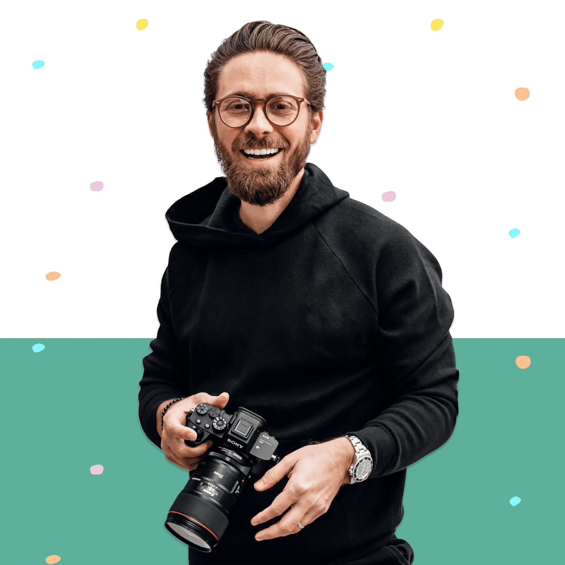 Bearded man in glasses and black hoodie holding a DSLR camera and smiling.