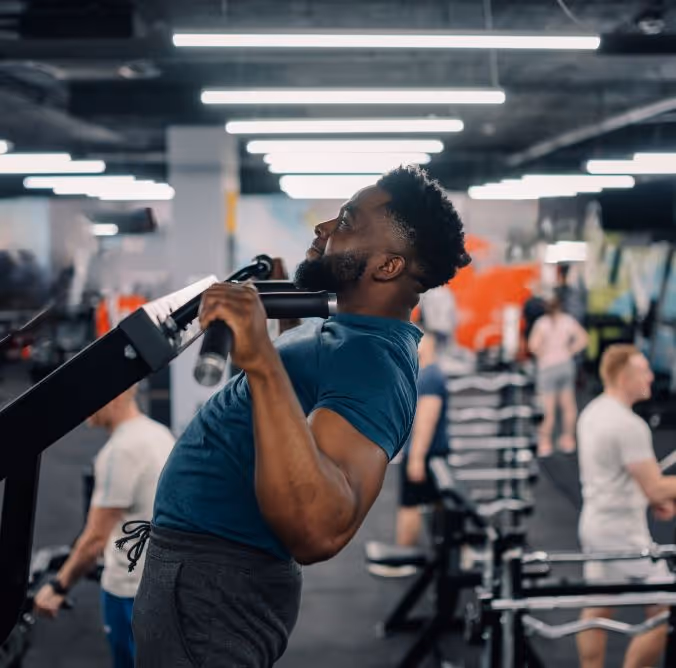 Man with beard doing pull-up exercise on a machine in a busy gym.