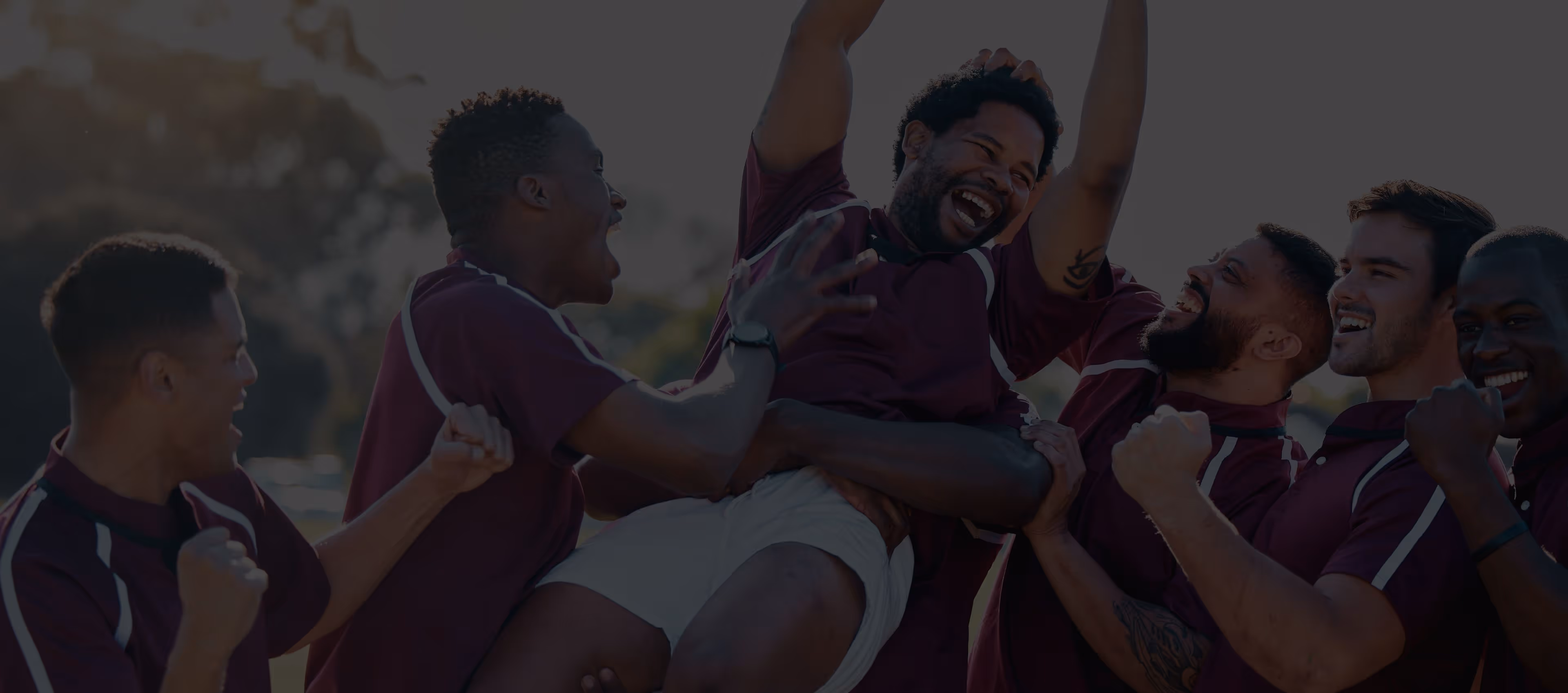 A group of men in matching maroon sports shirts cheerfully lift a teammate in celebration outdoors.