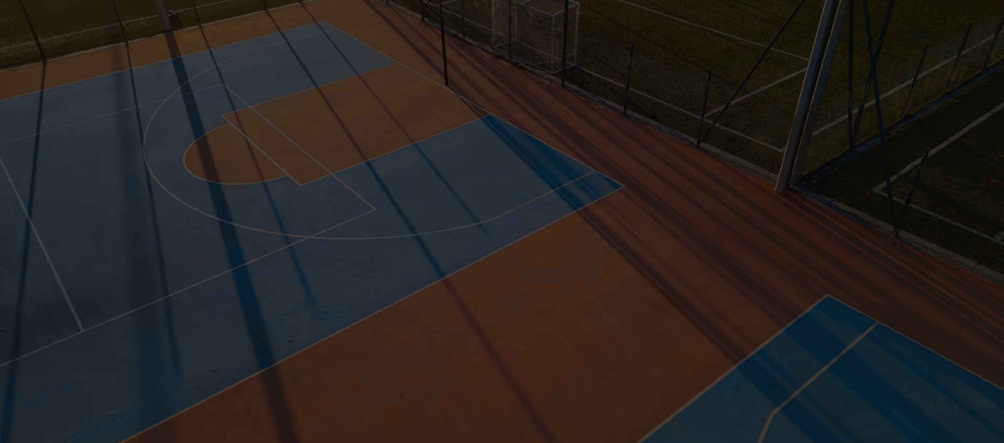 Empty outdoor basketball court with blue and orange surface under long shadows in late daylight.