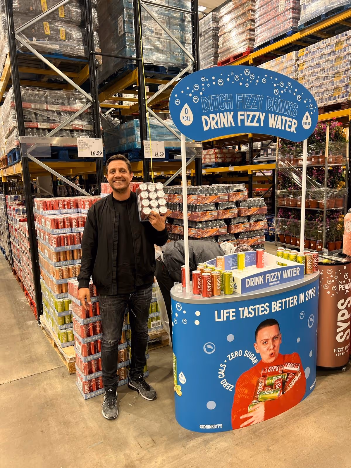 Man standing in warehouse store holding a pack of SYPS fizzy water cans next to promotional display booth.