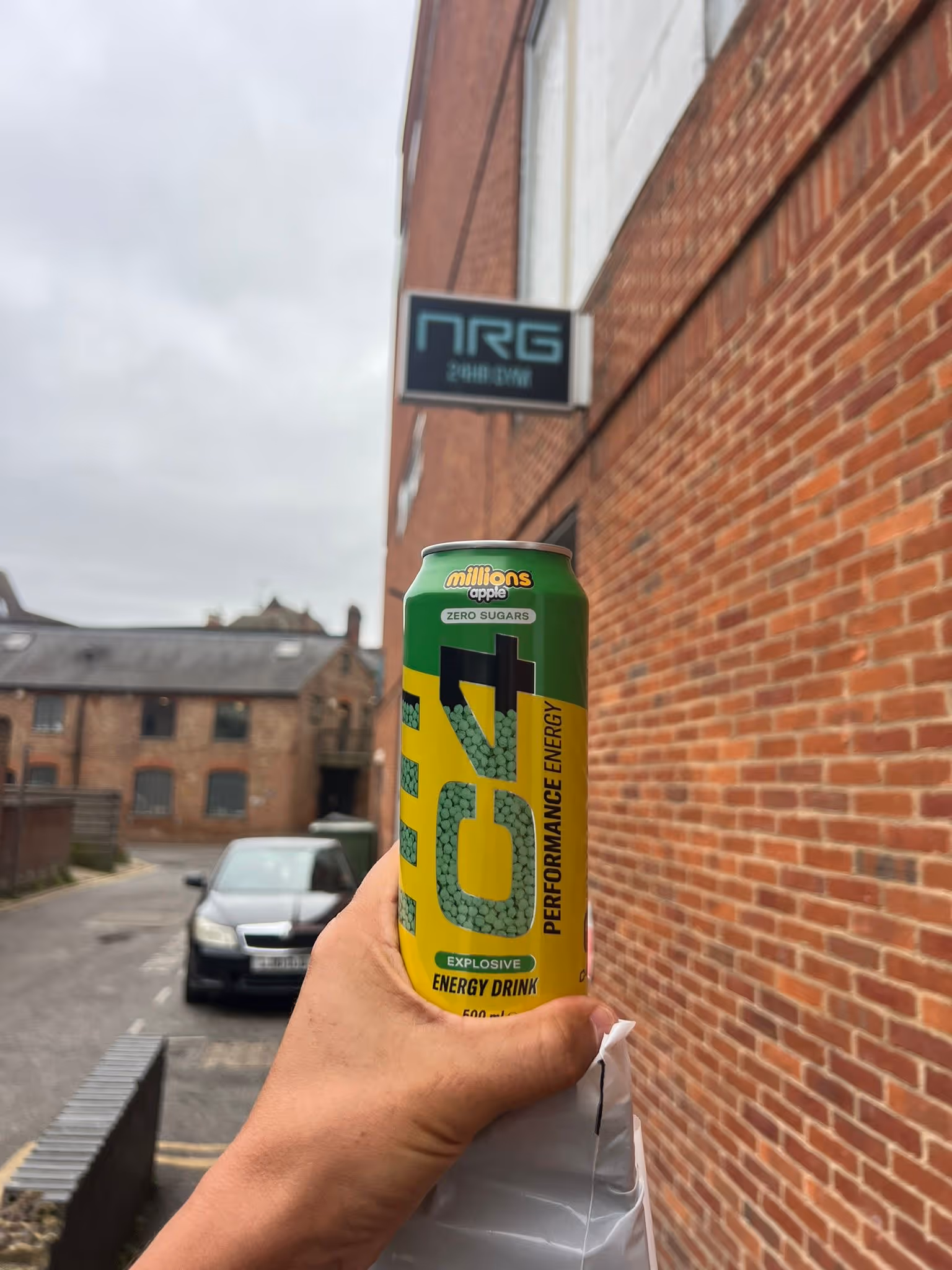 Hand holding a green and yellow C4 explosive energy drink can outdoors near brick buildings.