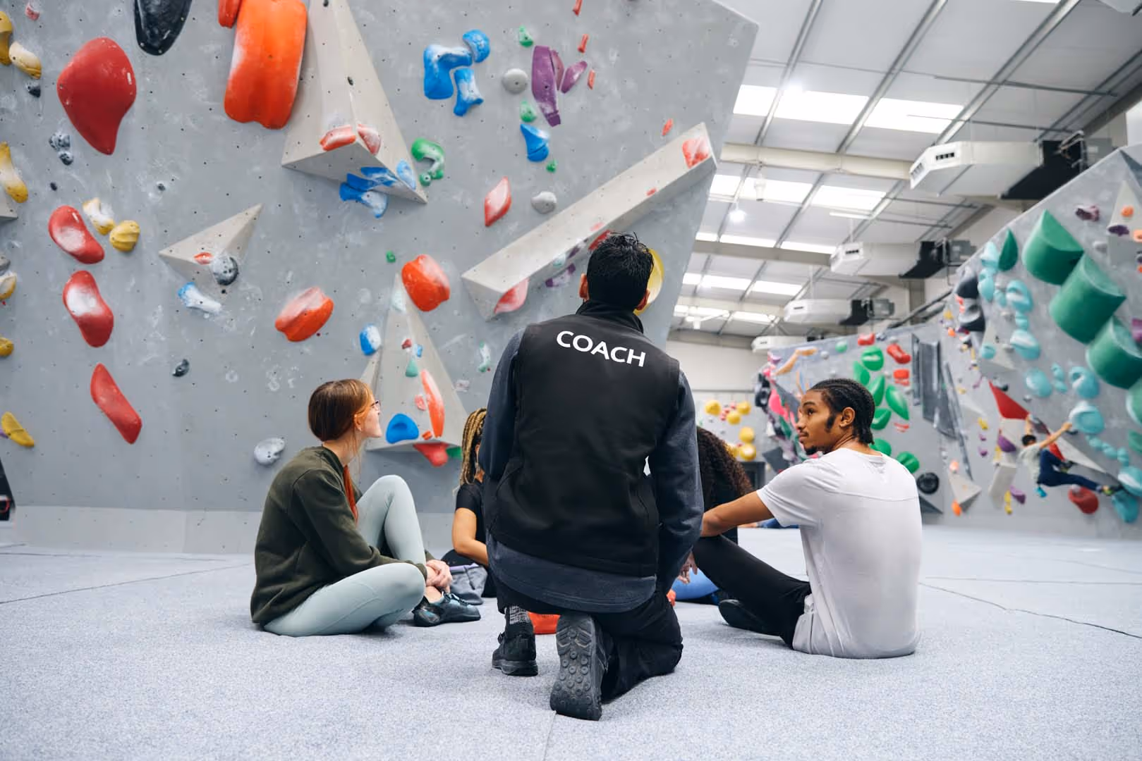 Coach kneeling and giving instructions to a group sitting on the floor in an indoor climbing gym.