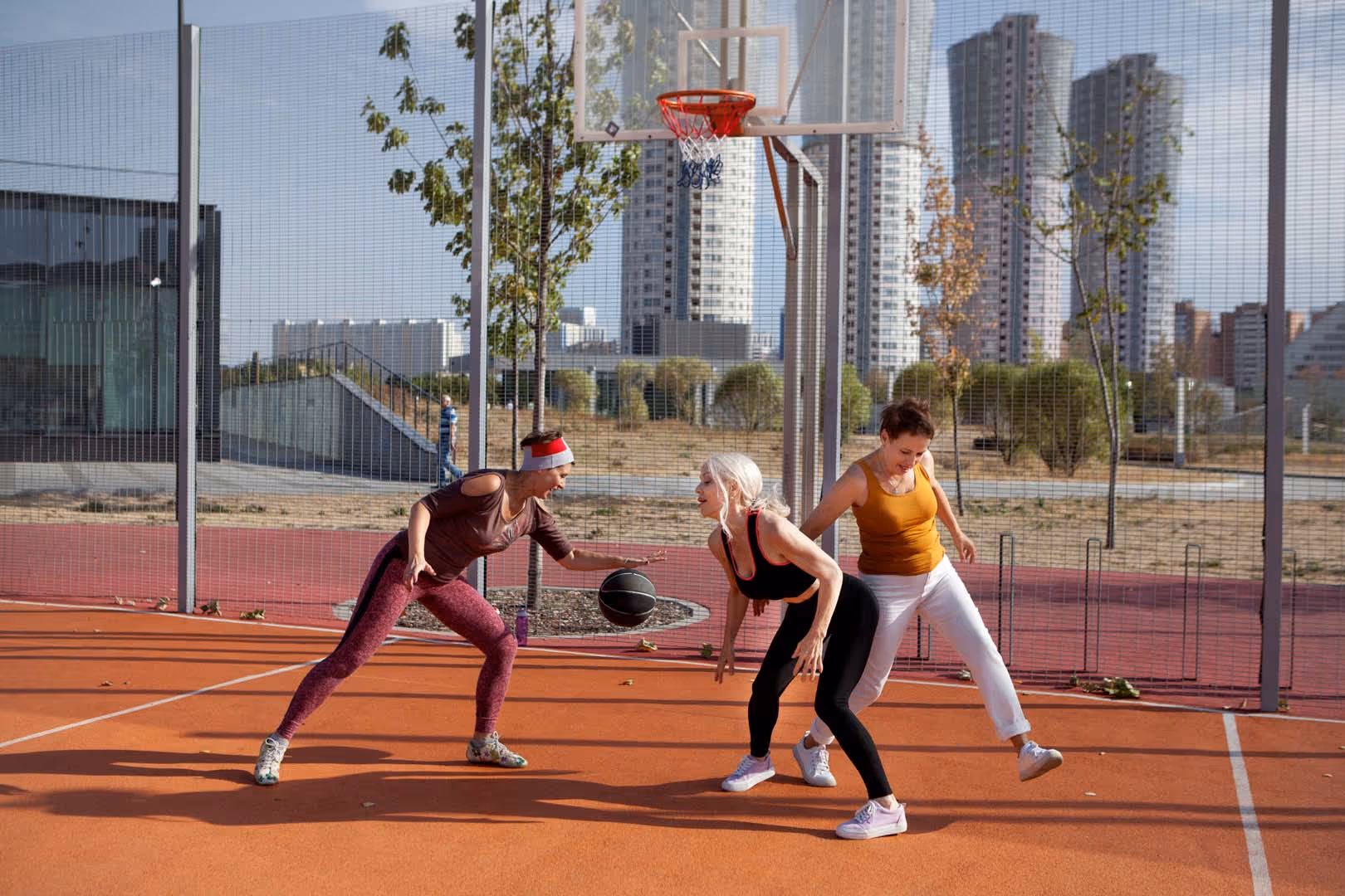 Three senior women playing basketball on an outdoor court in a city setting.