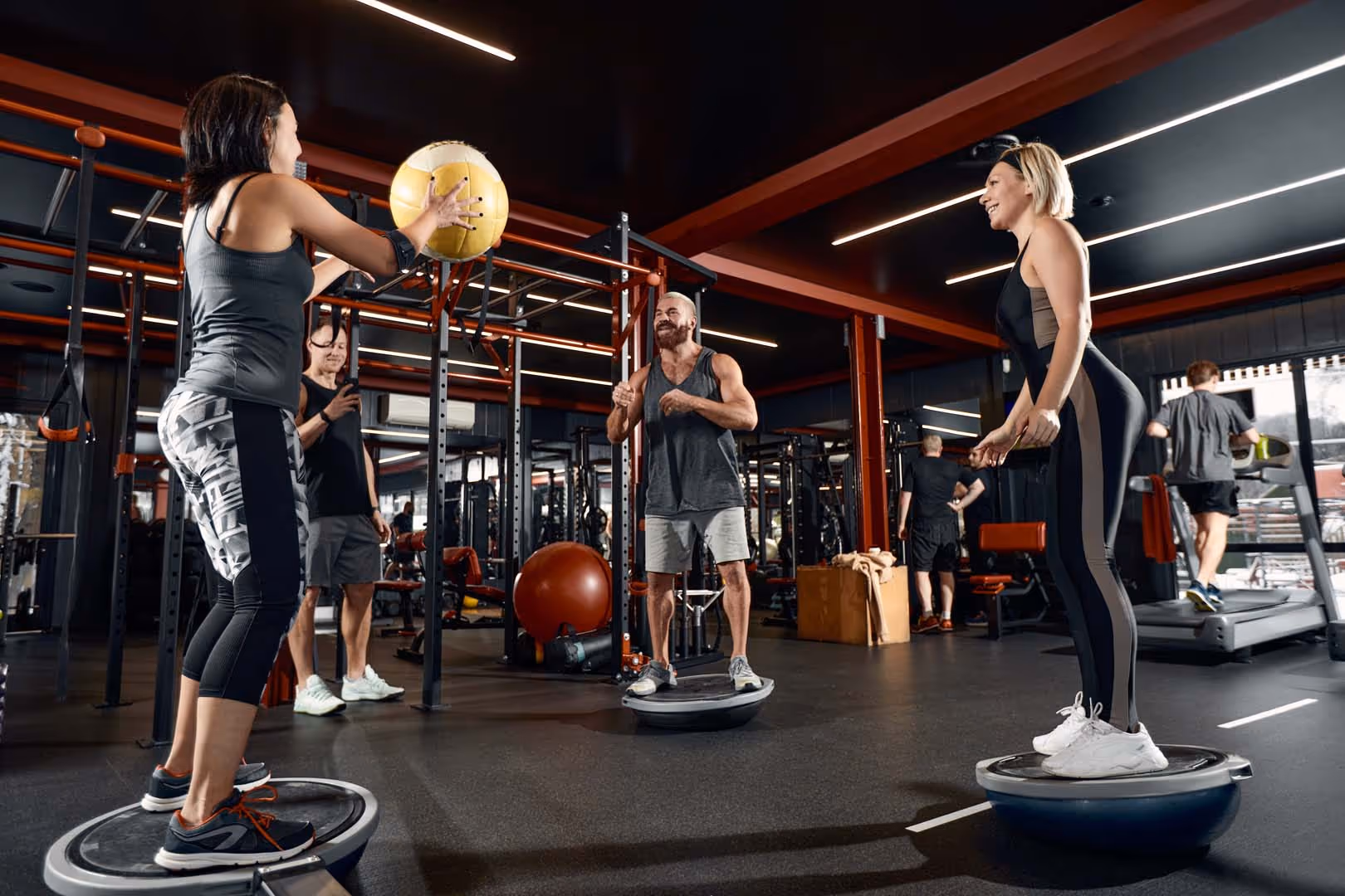 Three people balancing on bosu balls in gym, one catching a yellow medicine ball from another.