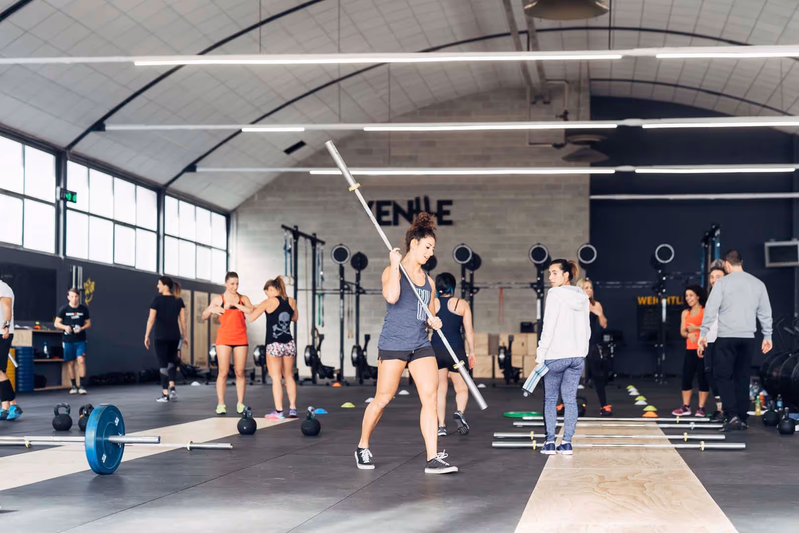 Group of people training with weights and barbells in a spacious gym with high ceilings.