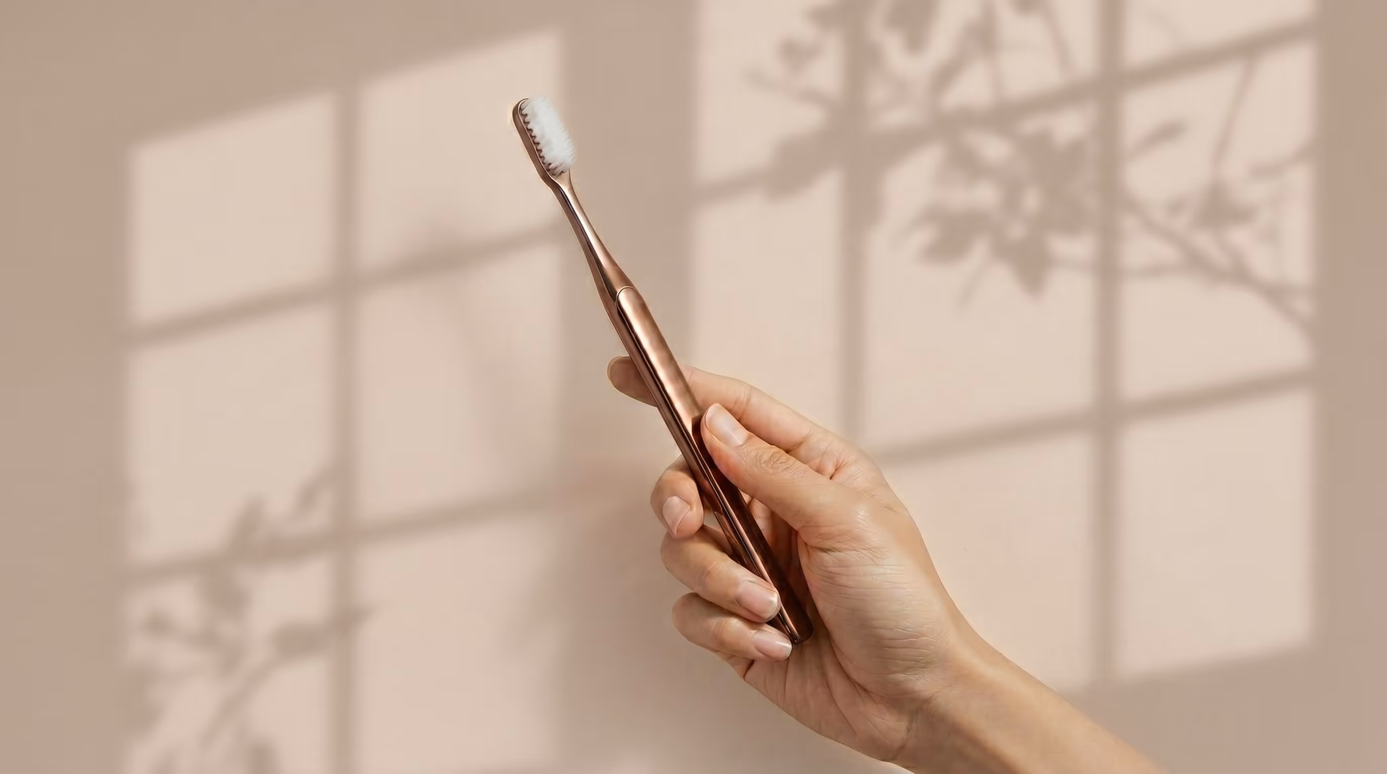 Hand holding a sleek metallic rose gold electric toothbrush with soft white bristles against a beige wall with window shadows and leaf silhouettes.