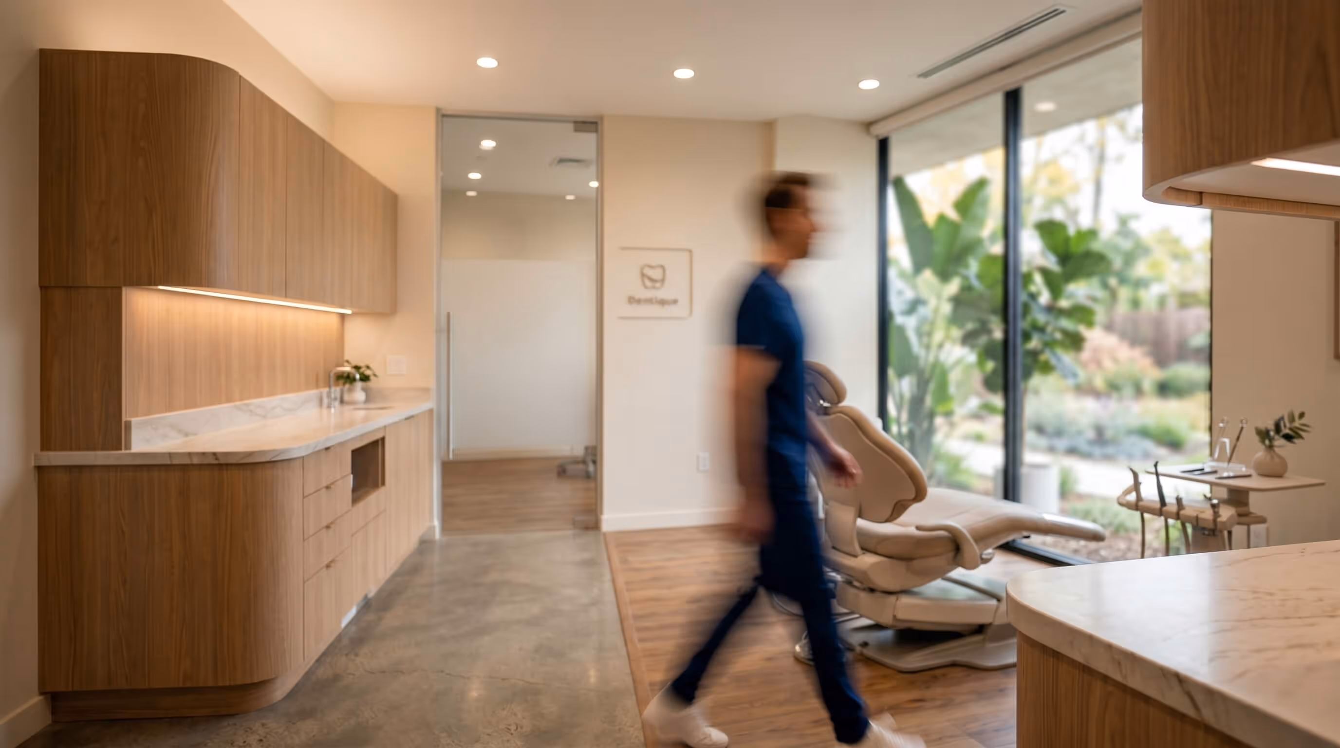 Blurred person walking in a modern dental office with wooden cabinets, beige dental chair, and large windows overlooking greenery.