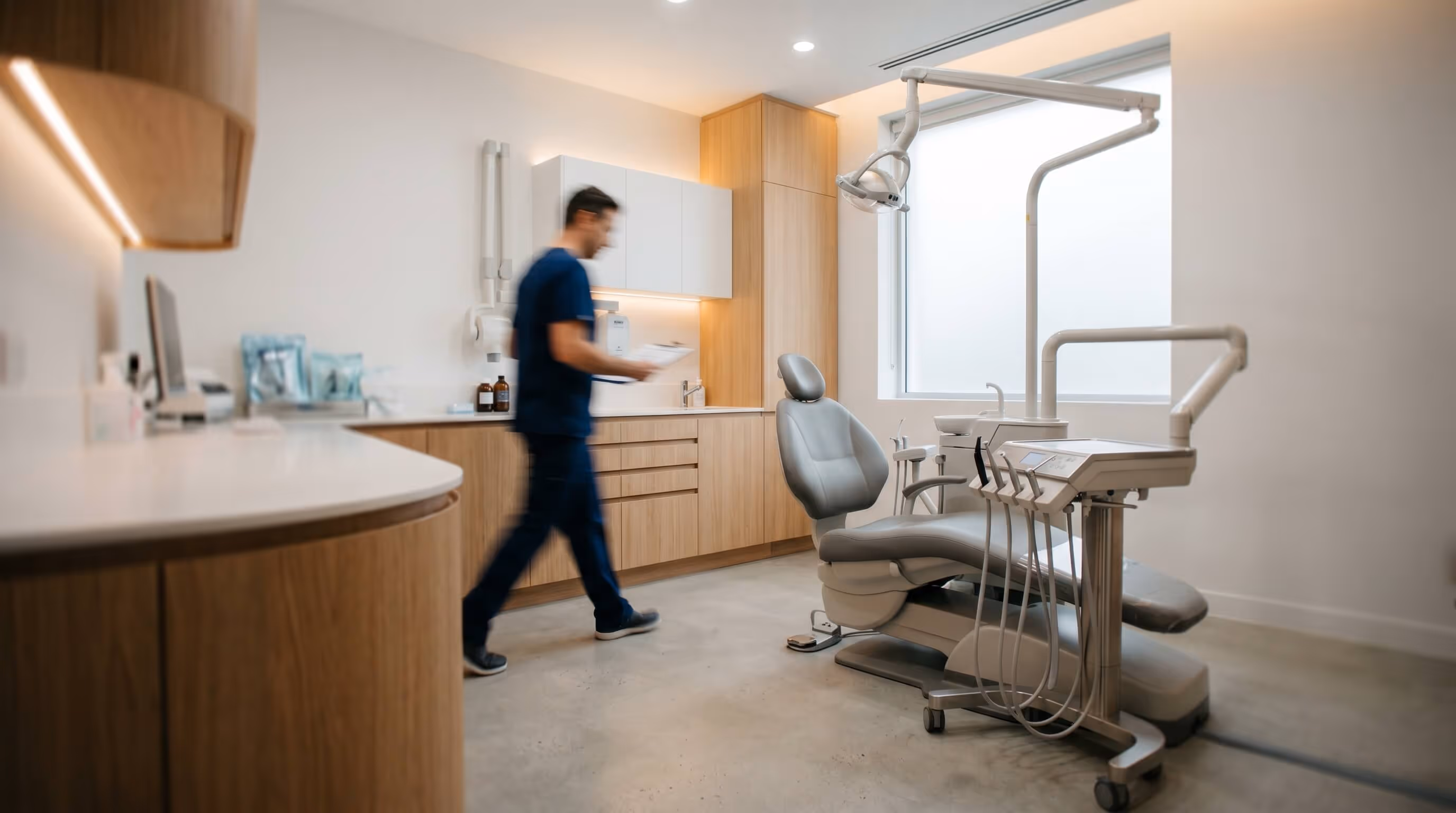 Modern dental clinic interior with a dentist walking near a gray dental chair and wooden cabinets.