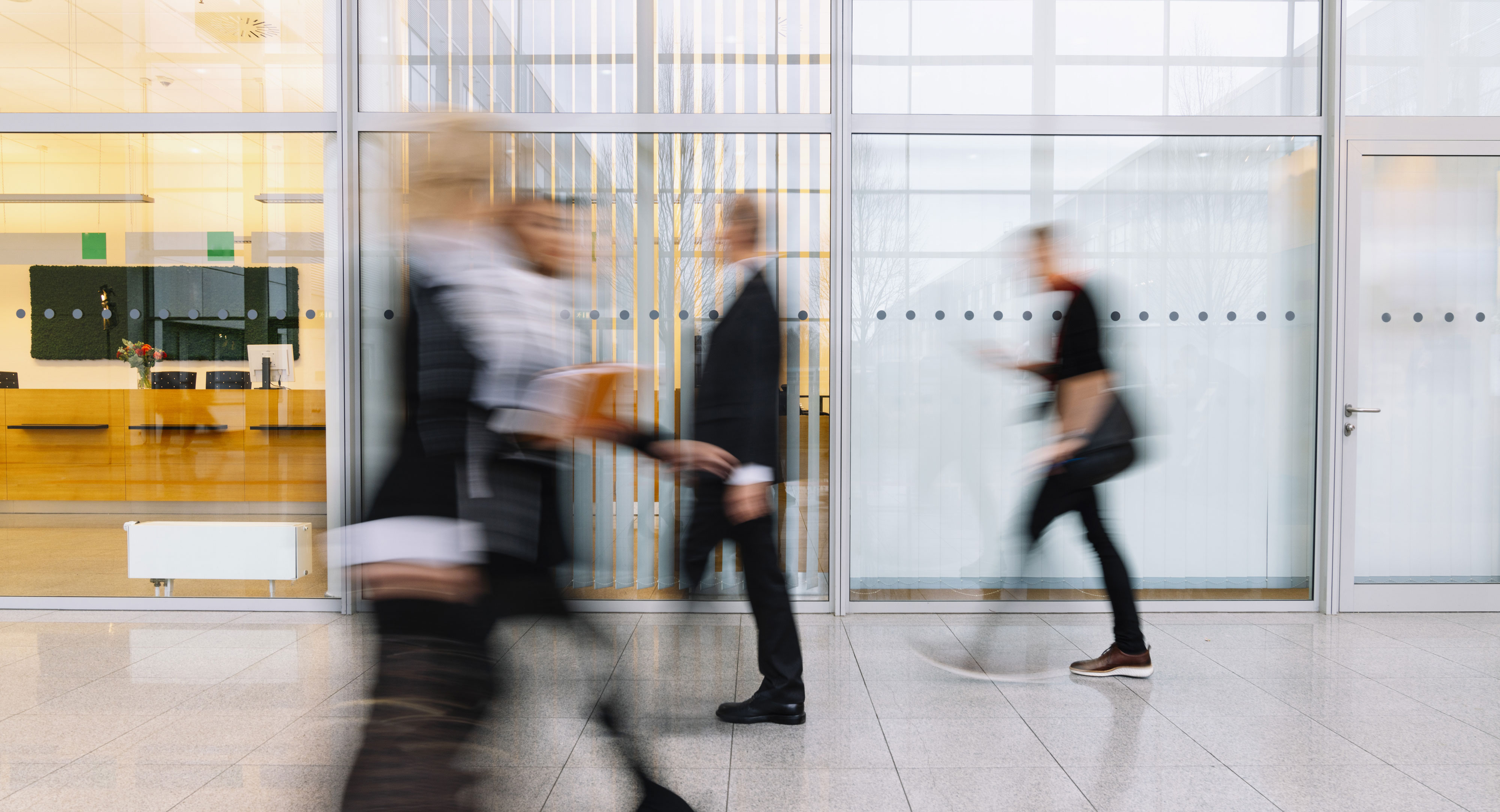 Blurred figures of people walking inside a modern office building with glass walls.