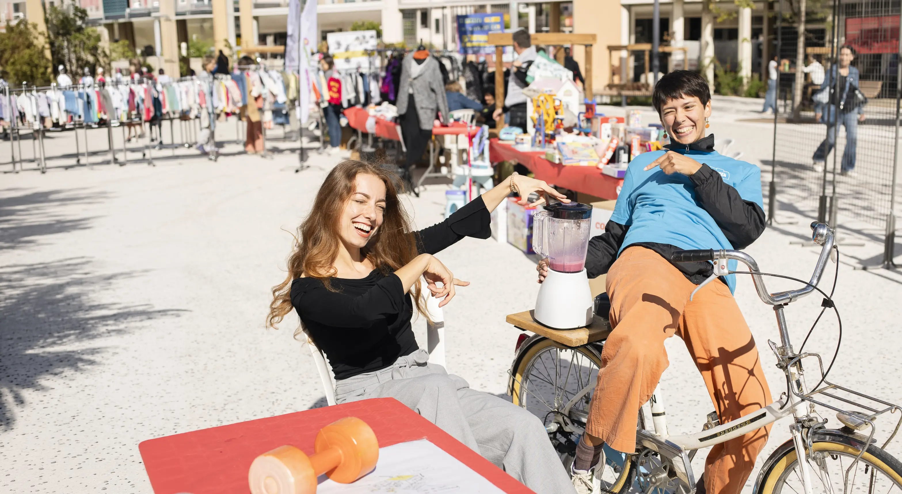 Deux femmes souriantes pointant un mixeur posé sur un vélo lors d'un marché en plein air.