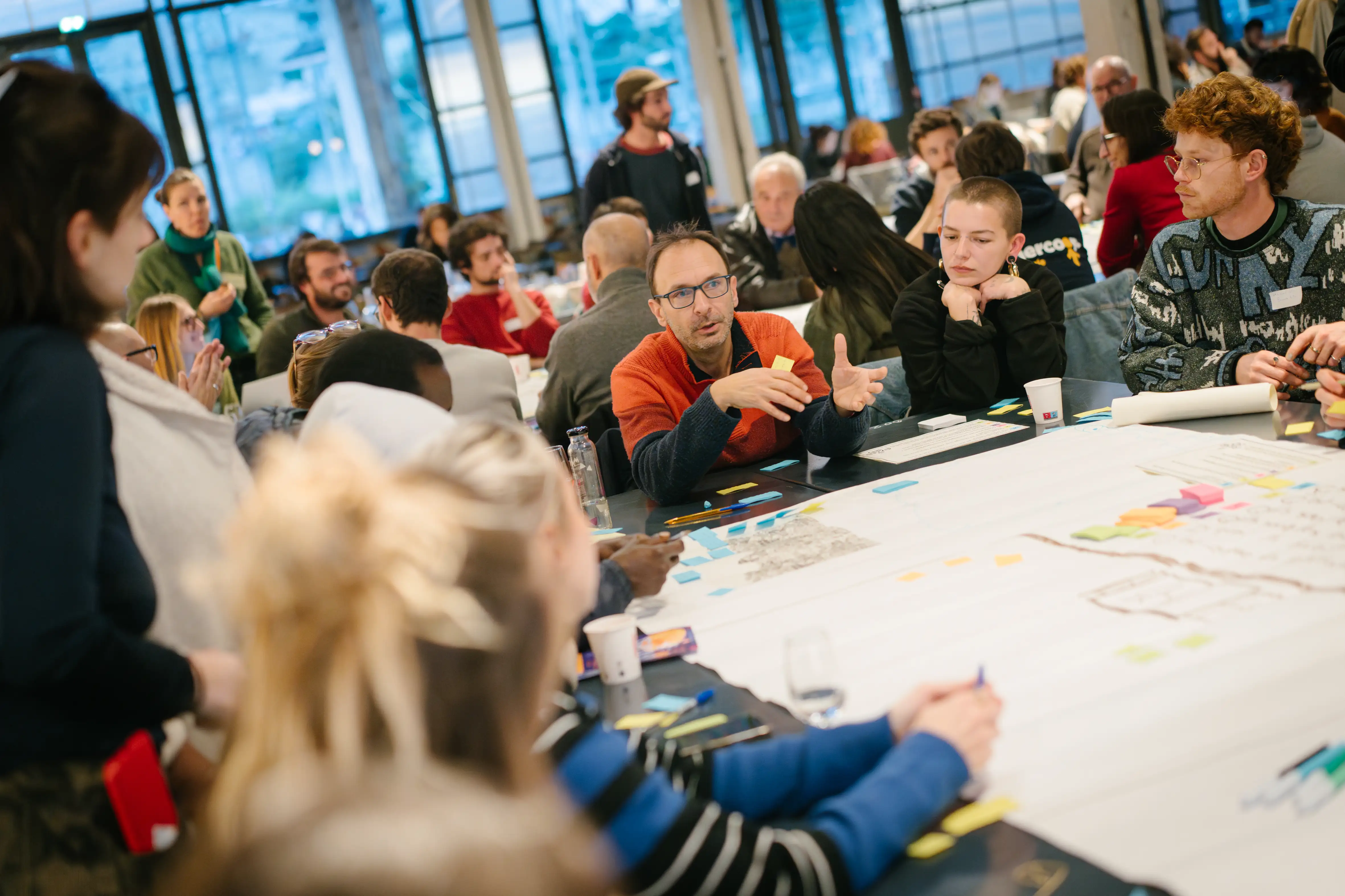 Groupe de personnes en réunion active autour d'une grande table avec post-its et documents.