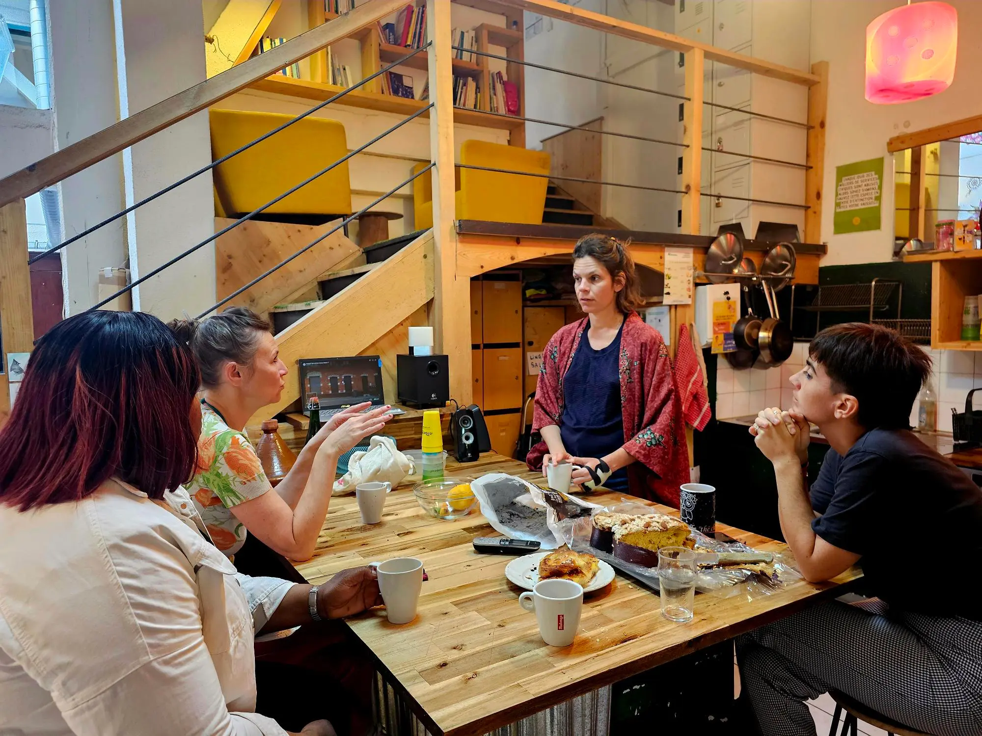 Quatre personnes discutent autour d'une table en bois avec boissons et pâtisseries dans une pièce conviviale.