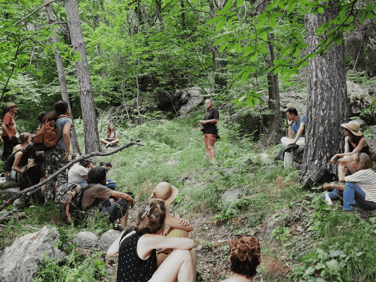 Un groupe de personnes assises dans une forêt écoutant un orateur debout parmi les arbres et rochers.