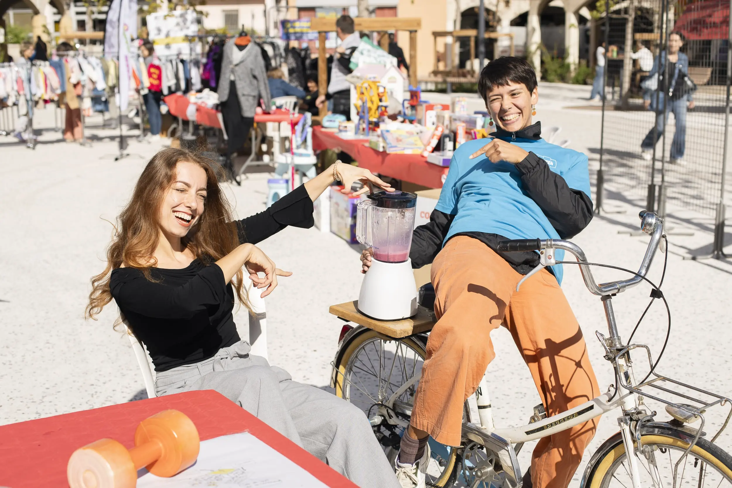 Deux femmes souriantes utilisent un vélo pour faire fonctionner un mixeur lors d'un marché en plein air.