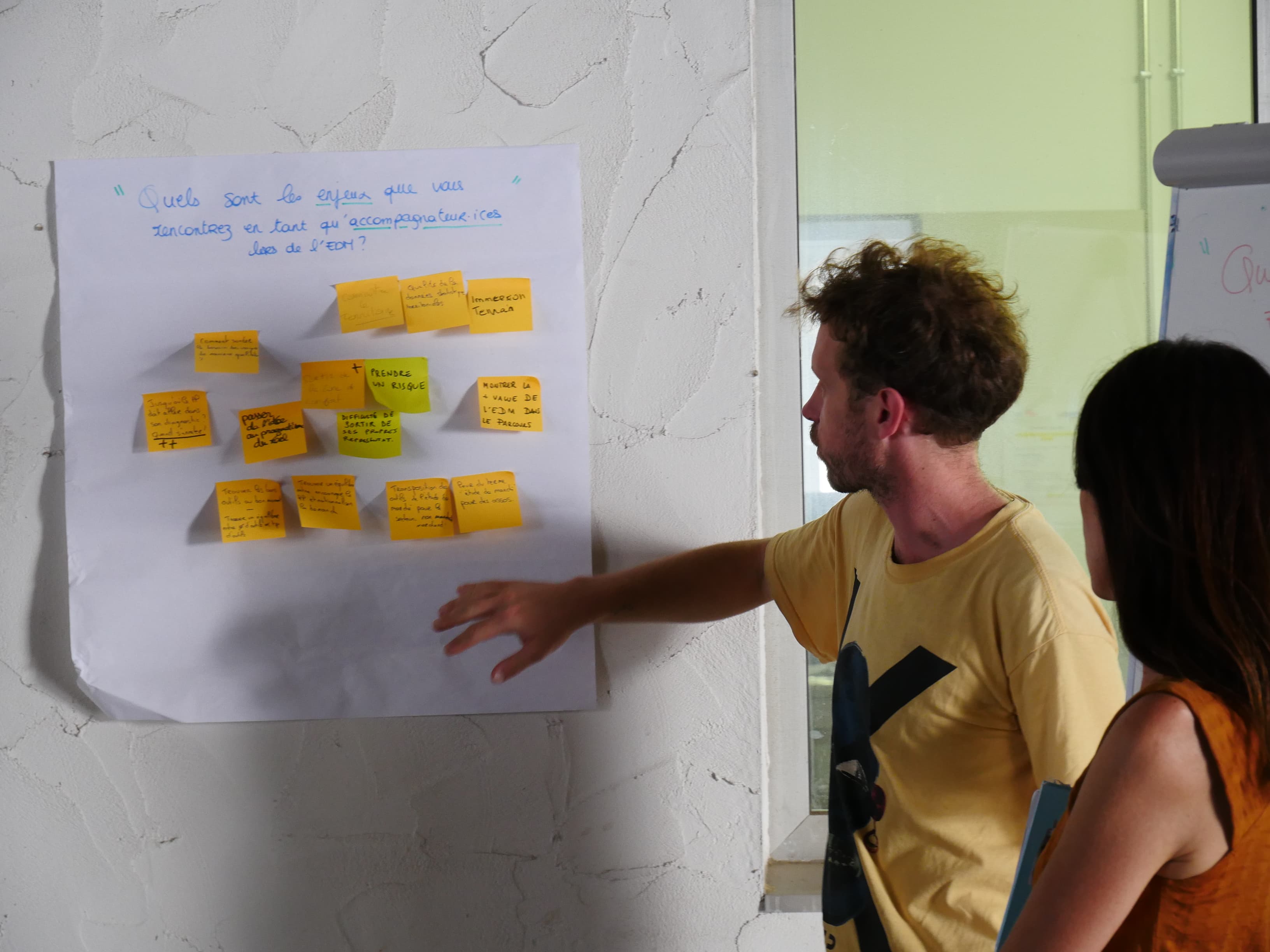 Deux personnes discutent devant un mur avec un poster et des post-its jaunes collés dessus.