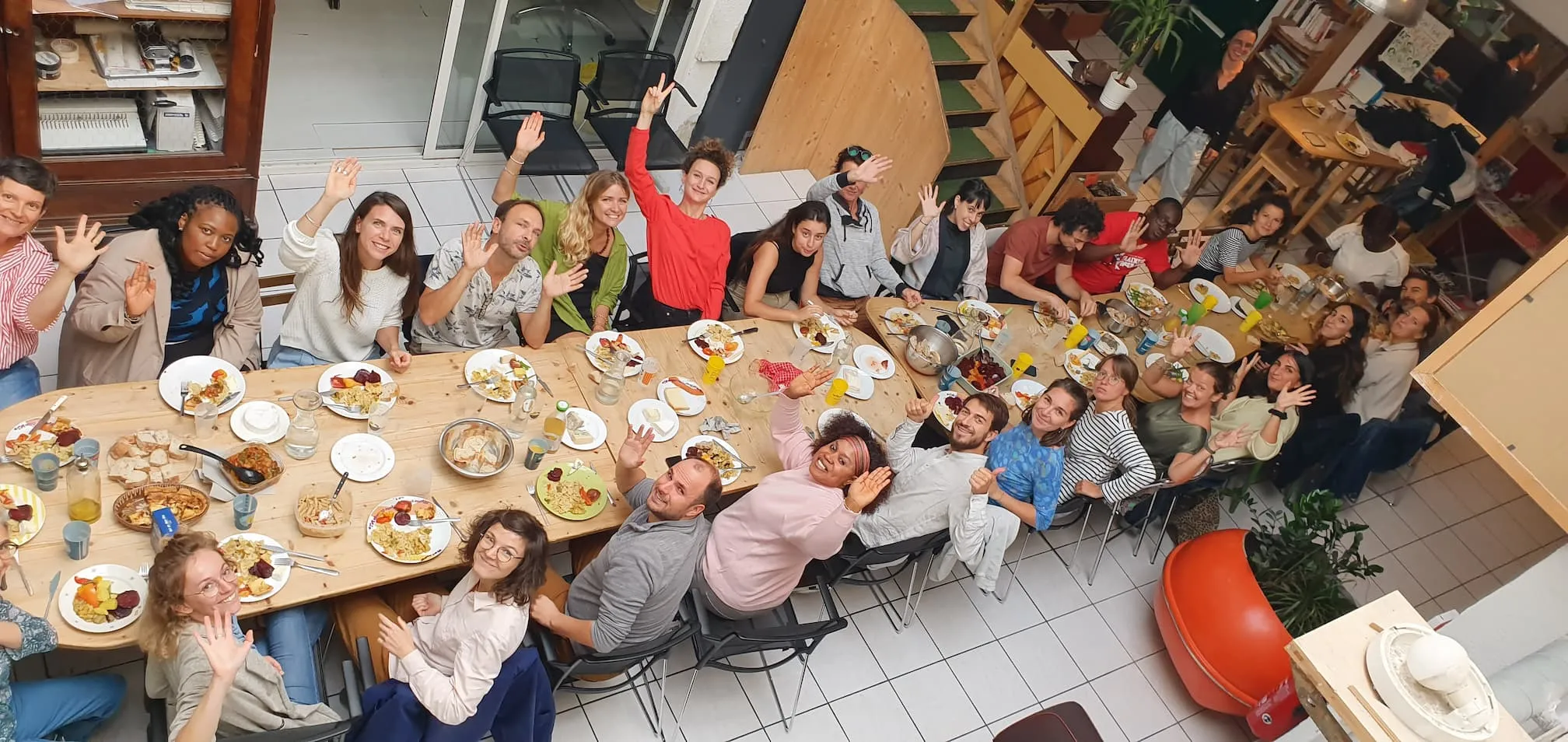 Groupe de personnes souriantes levant la main autour d’une table lors d’un repas convivial.