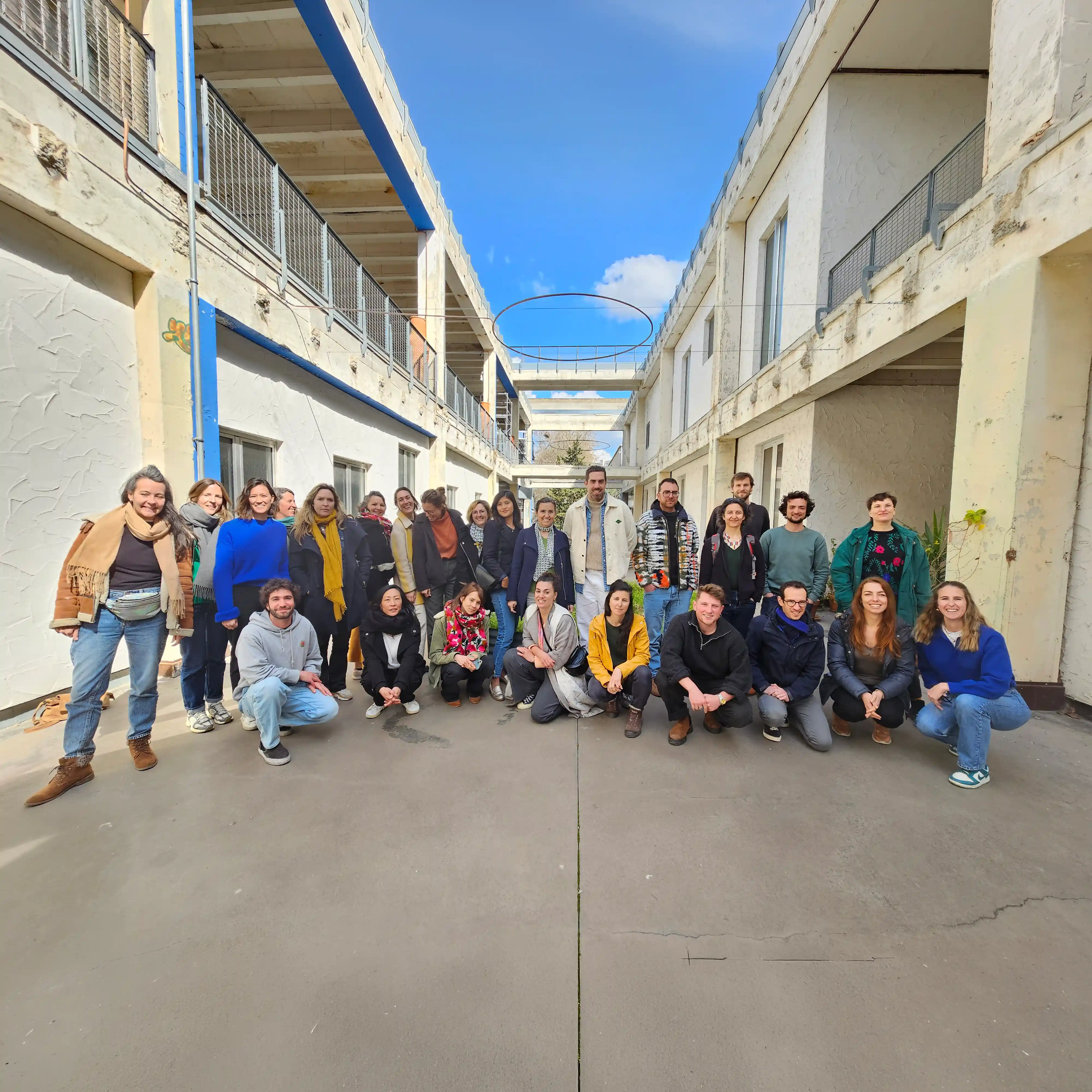 Groupe de 28 personnes posant dehors entre deux bâtiments sous un ciel bleu clair.