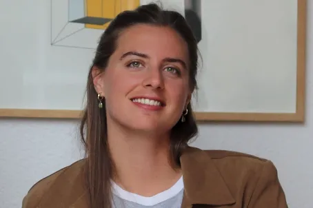 Jeune femme souriante avec des cheveux bruns, portant une veste marron claire et des boucles d'oreilles.