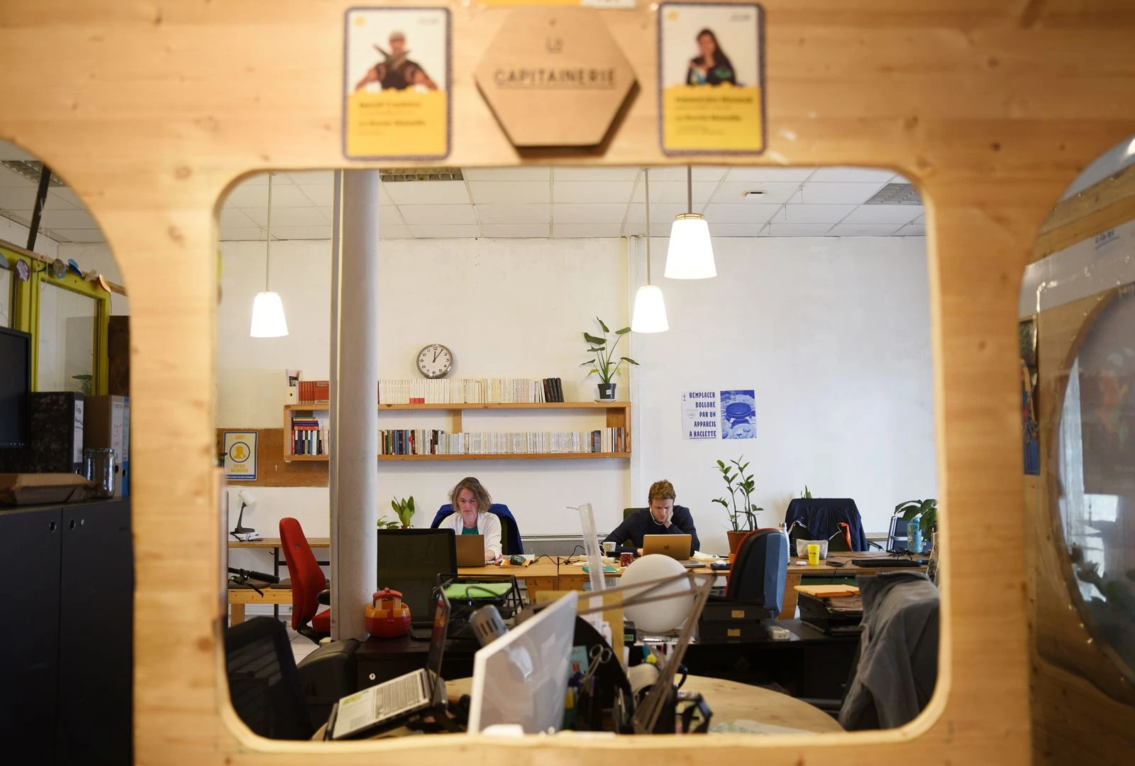 Deux personnes travaillent sur ordinateurs dans un espace de coworking lumineux avec plantes et étagères à livres.