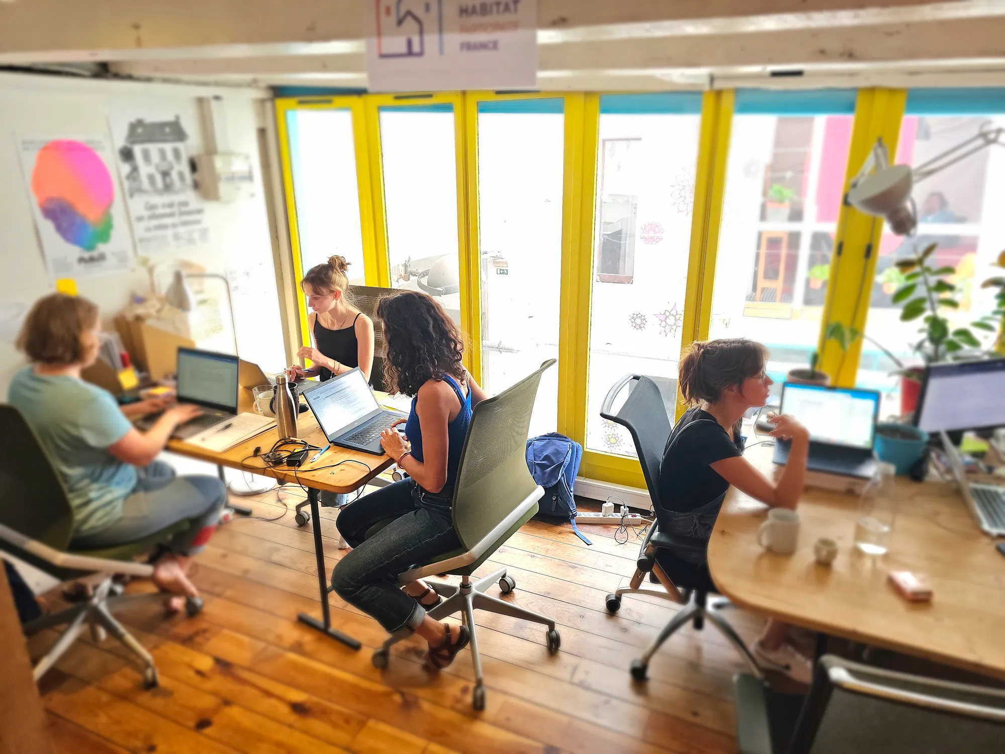 Quatre personnes travaillent sur des ordinateurs portables dans un espace de coworking lumineux avec fenêtres jaunes.