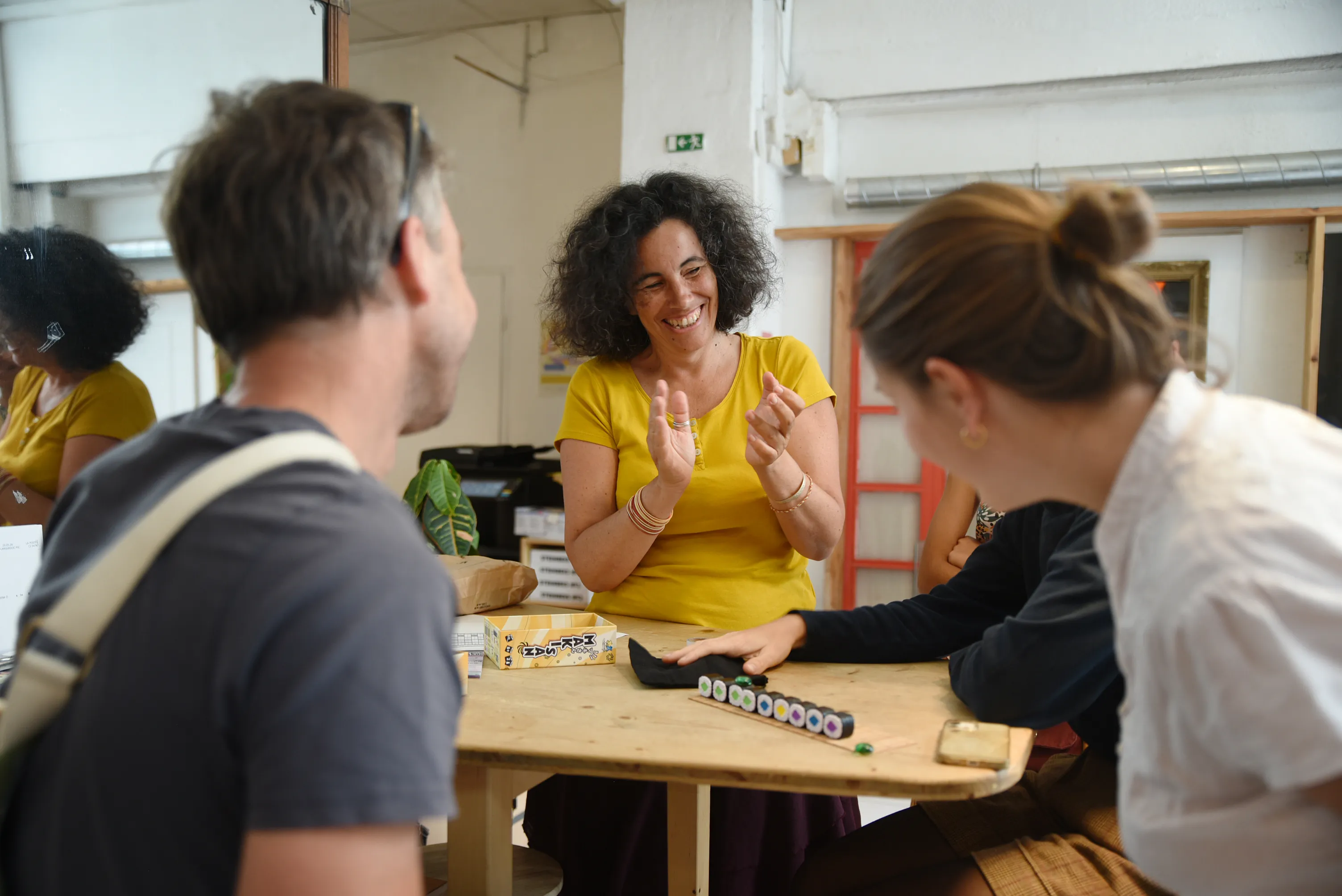 Trois adultes souriants jouent à un jeu de société autour d'une table en bois clair.