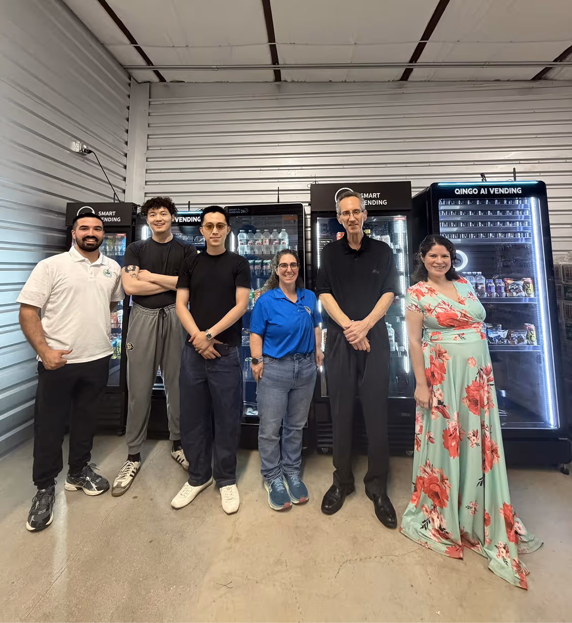 Group of six people standing in front of vending machines inside a warehouse setting.