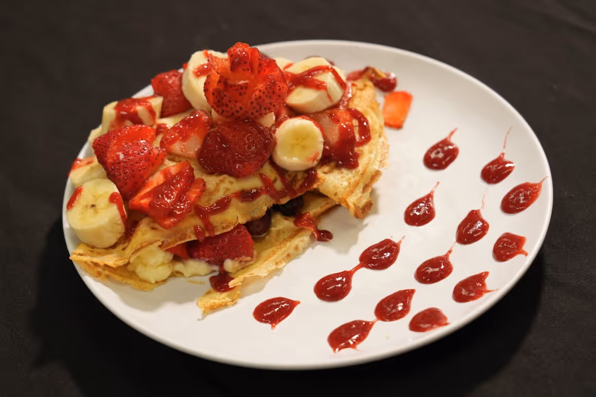 Crepes topped with banana slices, strawberries, and red sauce with dots of sauce on the plate.