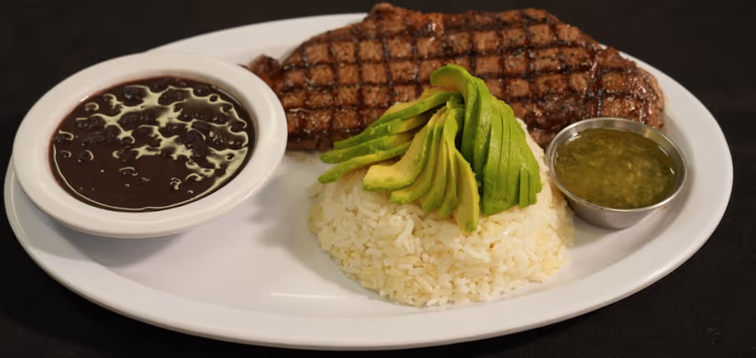 Grilled steak with sliced avocado atop white rice, served with black beans and green sauce on a white plate.