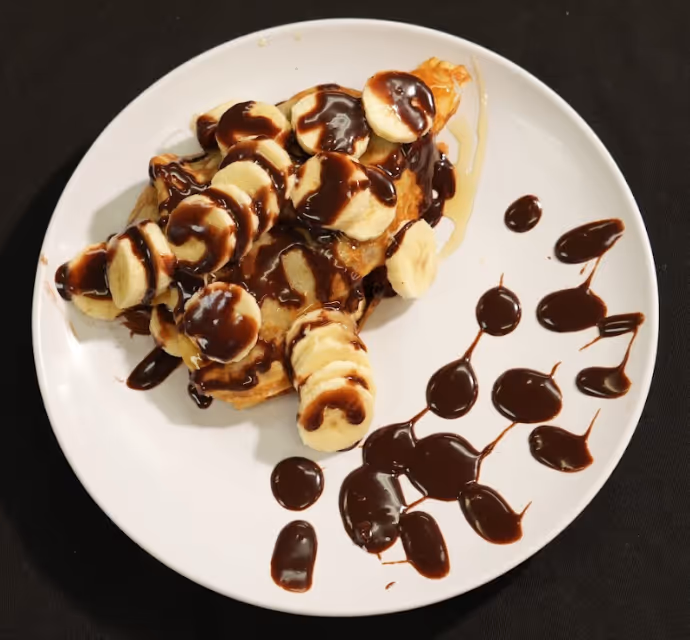 Plate of crepes topped with banana slices and drizzled with chocolate sauce, with extra chocolate sauce on the side.