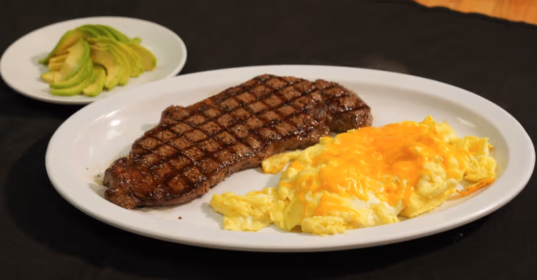 Grilled steak with scrambled eggs topped with melted cheddar cheese and a side plate of sliced avocado.