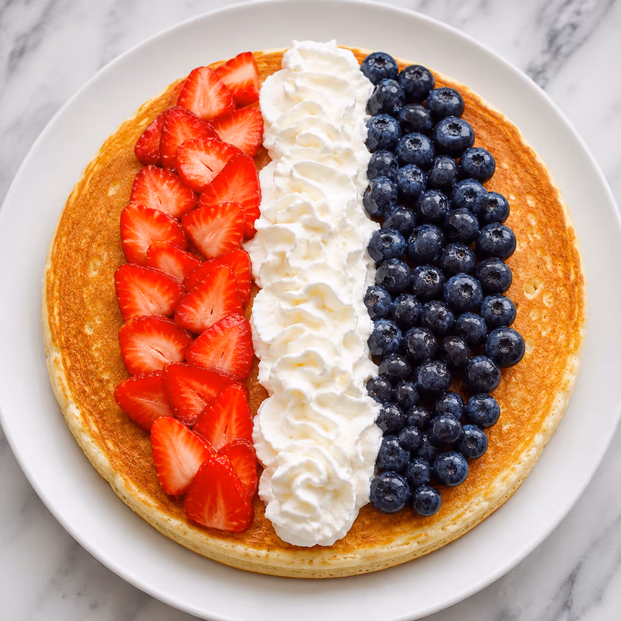 Pancake topped with rows of sliced strawberries, whipped cream, and blueberries on a white plate.
