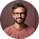 Smiling man with dark curly hair, beard, glasses, and a brown shirt against a dark background.