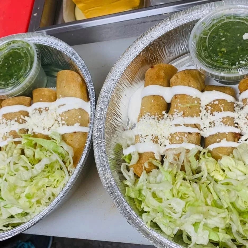 Two foil containers with rolled tacos topped with white sauce and cheese, shredded lettuce, and a small cup of green salsa.