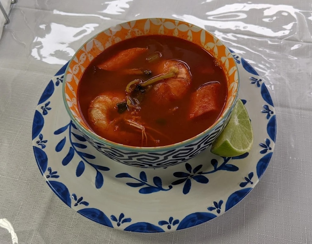 Bowl of red shrimp soup with vegetables served with a lime wedge on a blue and white plate.