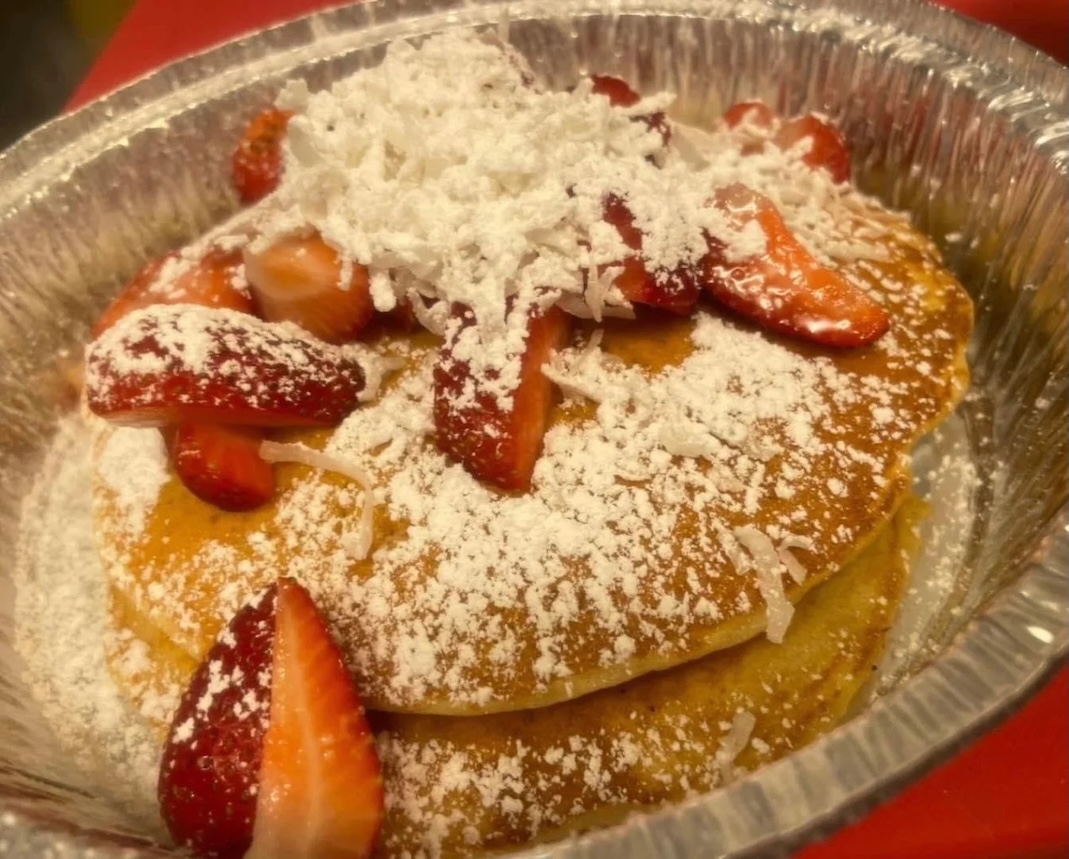 Two pancakes topped with sliced strawberries and powdered sugar in a foil container.