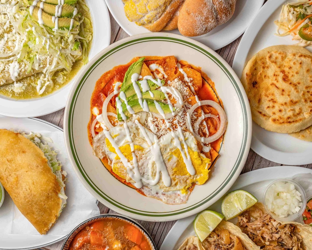 Plate of chilaquiles topped with fried eggs, avocado slices, onion rings, cheese, and crema sauce among Mexican dishes.