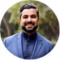Smiling man with beard and dark hair wearing a blue jacket outdoors with blurred background.