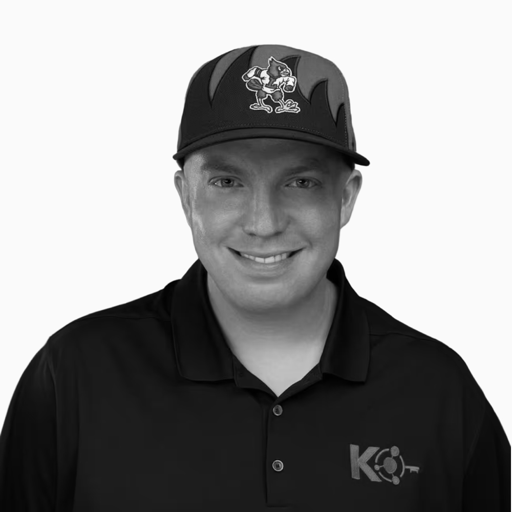 Professional black and white headshot of Garret, Founder at Key Outreach, smiling while wearing a black polo shirt with the Key Outreach logo and a black baseball cap featuring a cartoon bird mascot, isolated on a light gray background.